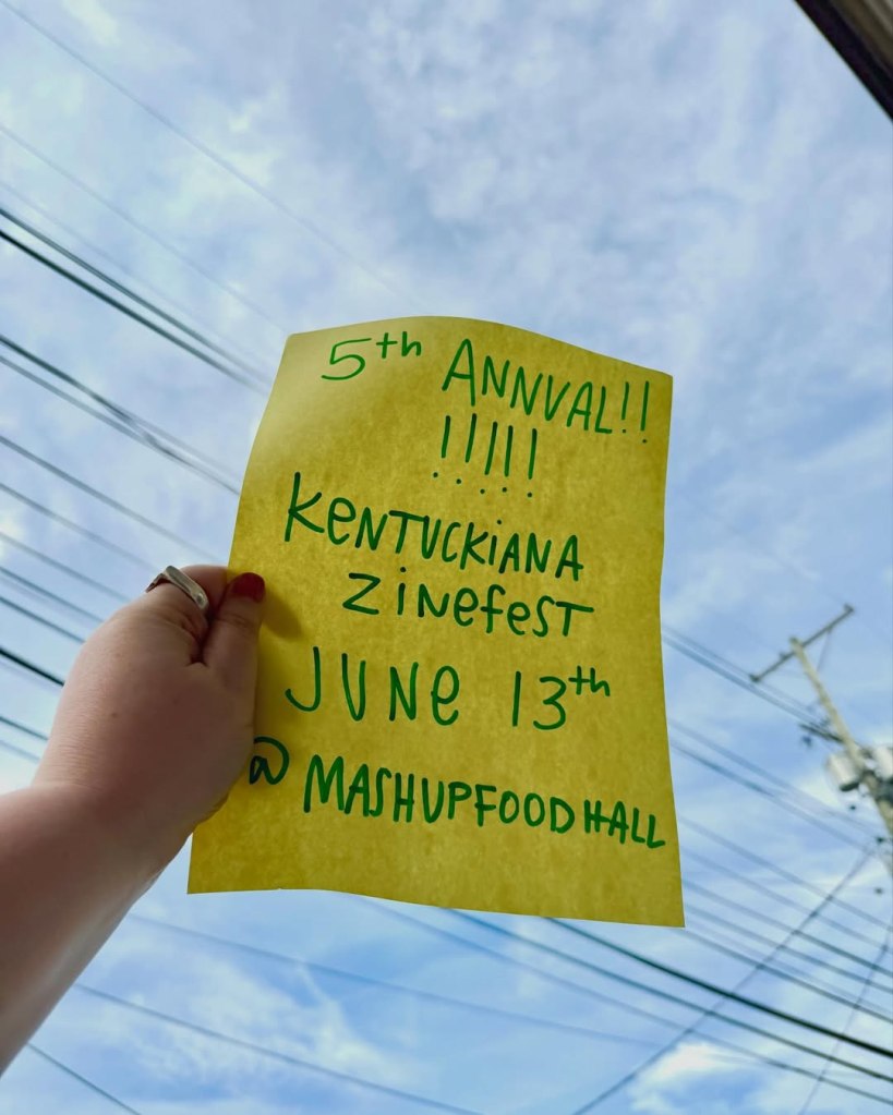 A hand holding a yellow flyer to the sky with blue handwritten text about the Zine Fest.